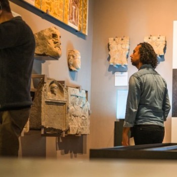 Young Caucasian woman reading through a digital tablet with data of the displayed objects while her biracial husband is checking out the stone remains of an Ancient Roman town. Candid shot over the display cases in the middle of museum gallery.